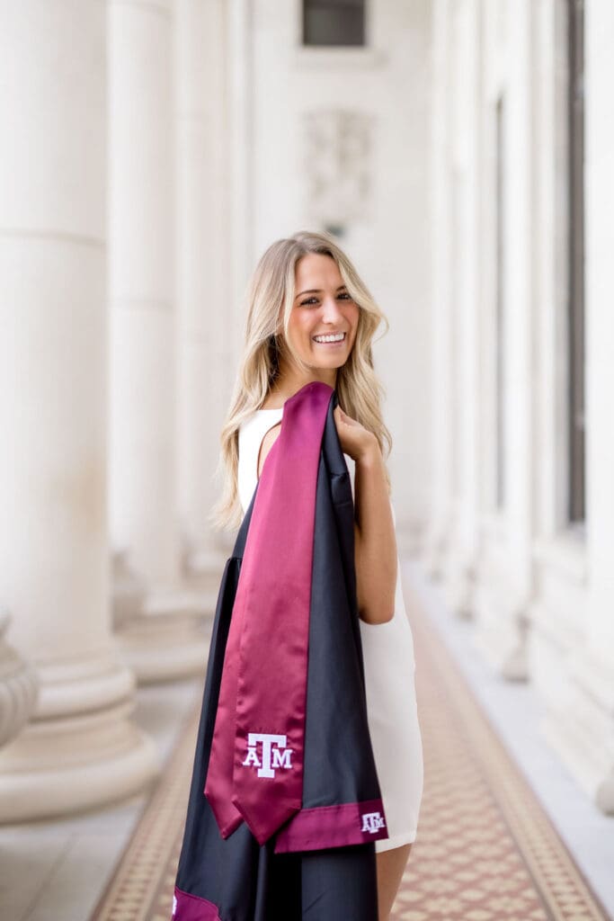Captured by a Texas A&M Photographer in College Station, Adeline in a white dress with maroon stole standing at the iconic Administration Building on campus for her graduation portraits.