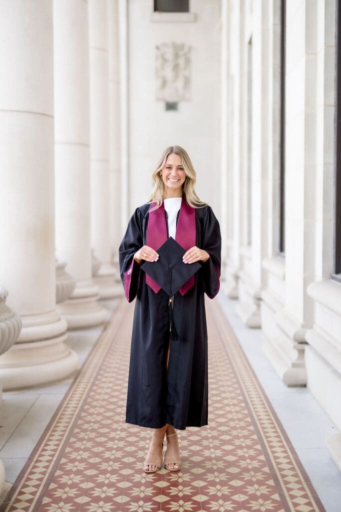 Captured by a Texas A&M Photographer in College Station, Adeline in a white dress with maroon stole standing at the iconic Administration Building on campus for her graduation portraits.